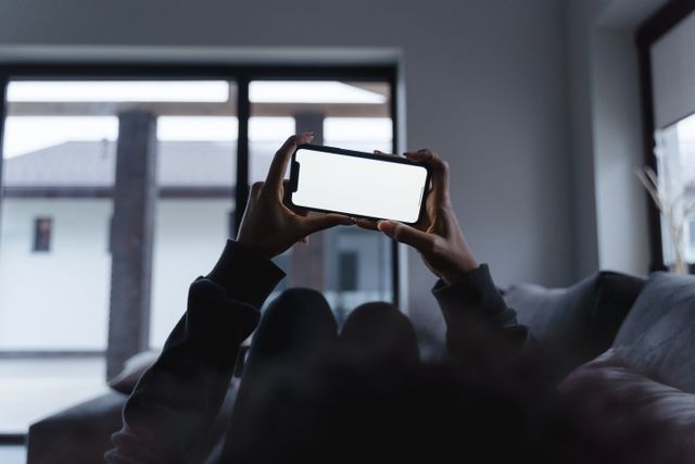 Ruke drže mobilni telefon vodoravno sa belim ekranom ispred prozora